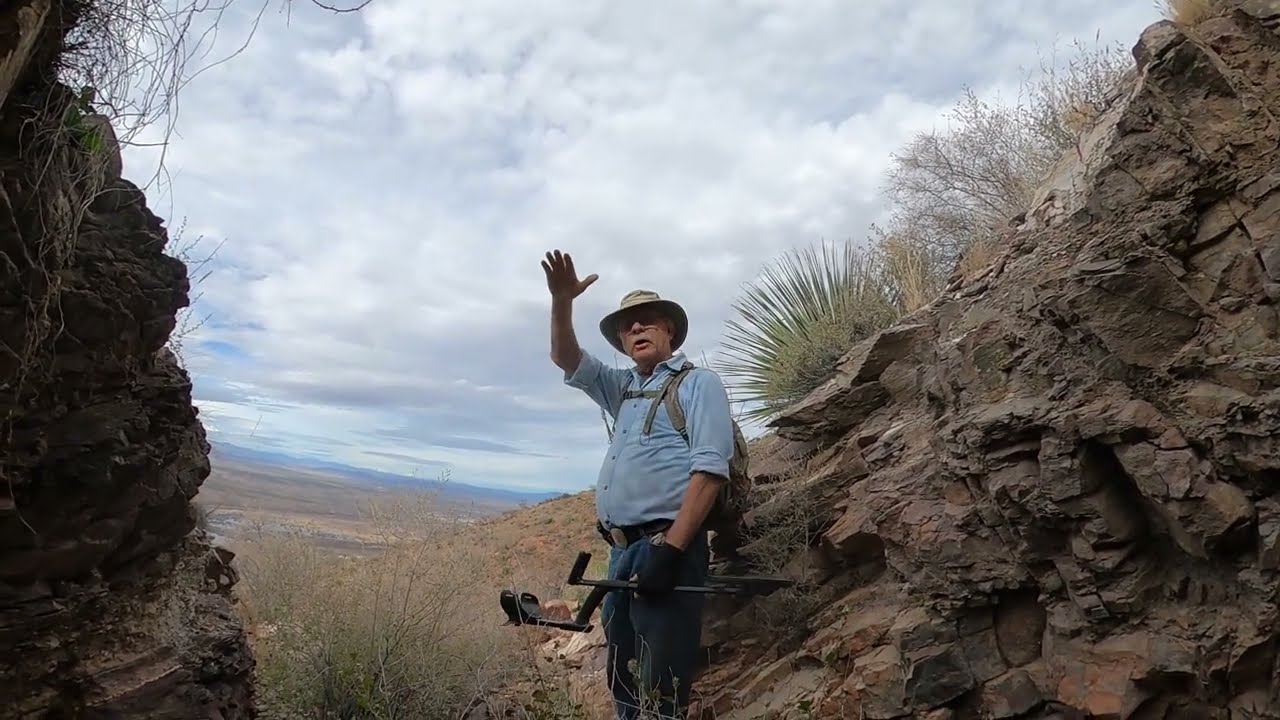 A man wearing a wide-brimmed hat, blue shirt, and backpack stands between rocky cliffs in a desert landscape, raising one hand while holding a metal detector in the other. The sky above is partly cloudy.