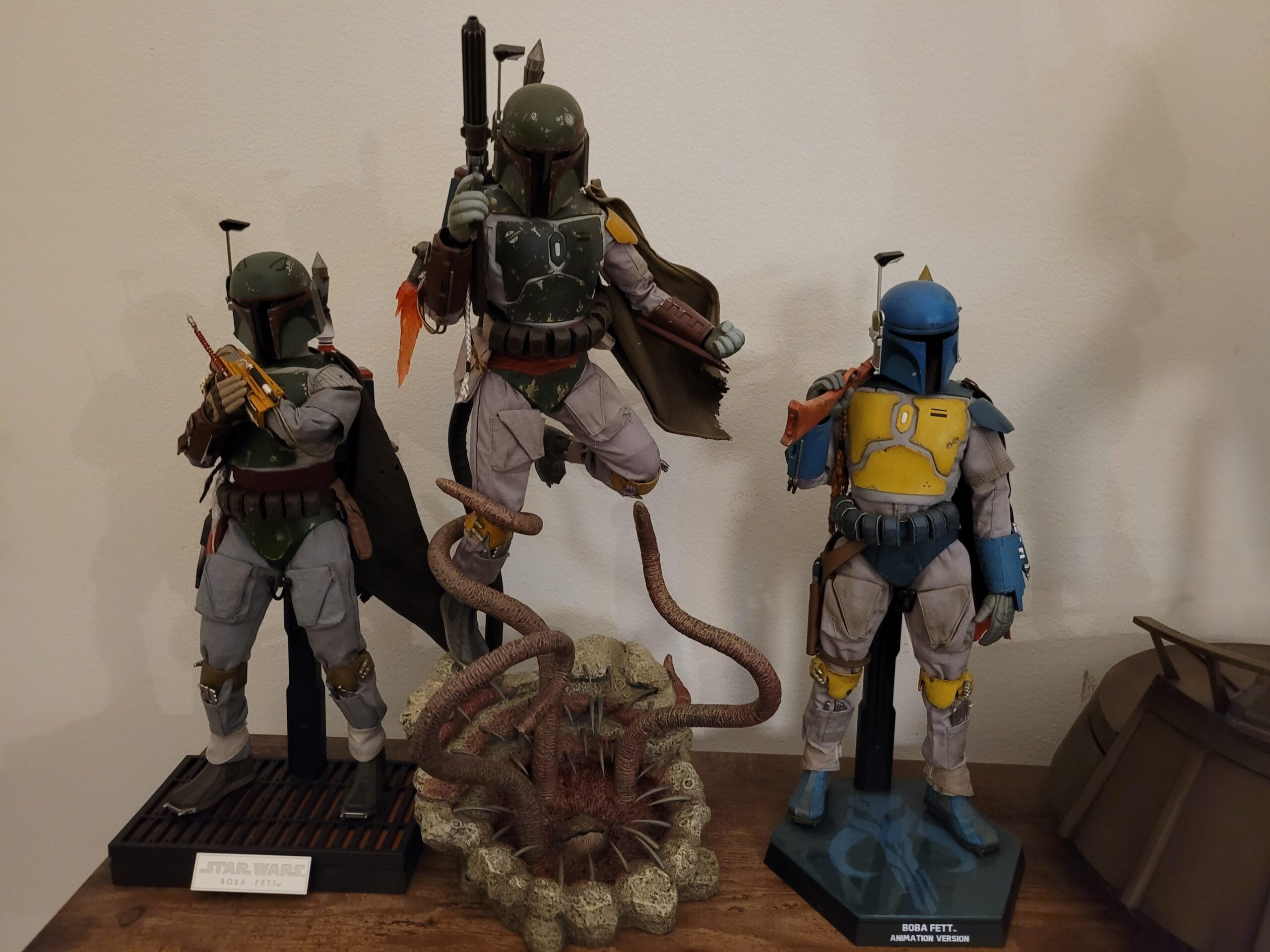 Three detailed Boba Fett action figures are displayed on a wooden surface. The center figure is elevated above a Sarlacc pit model, while the other two figures stand on either side in different armor colors and poses.