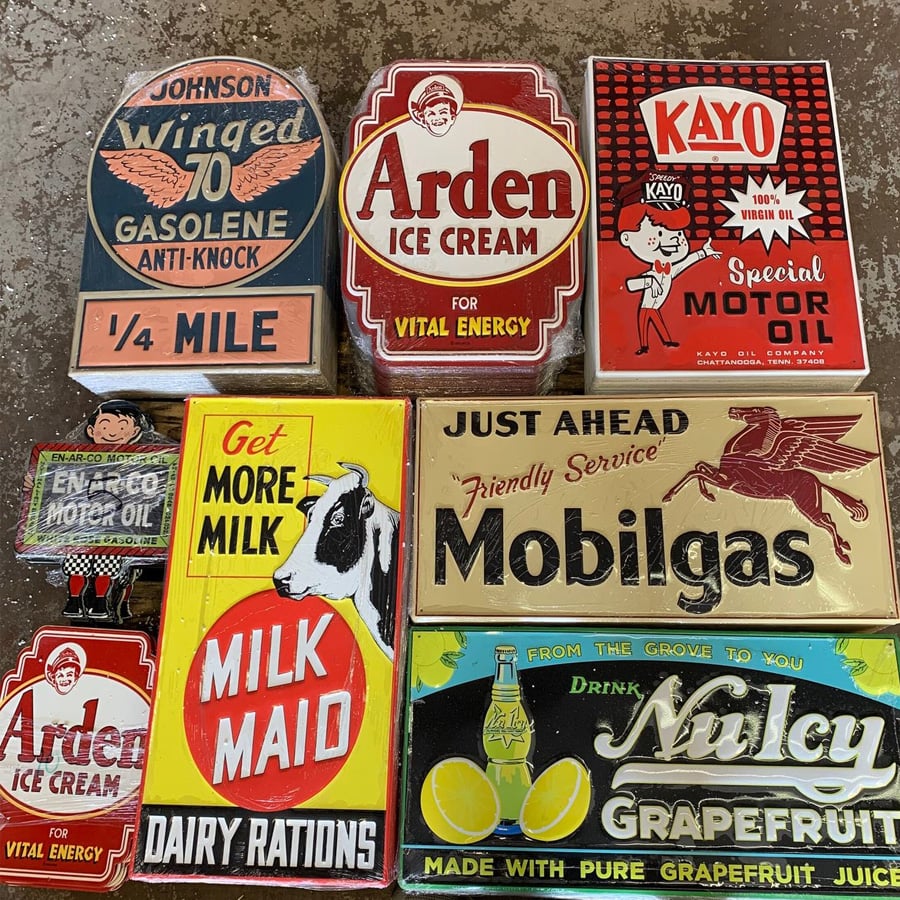 A collection of vintage-style metal signs advertising gasoline, motor oil, ice cream, milk, and grapefruit juice, arranged on a textured surface. Each sign features bold, retro fonts and colorful graphics.