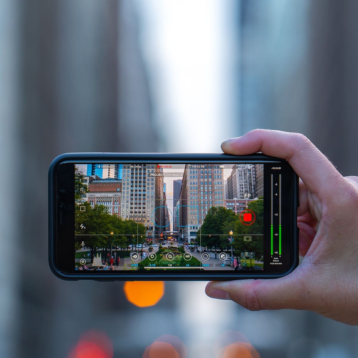 A hand holds a smartphone horizontally, capturing a photo of tall buildings and a street scene with trees and people. The phone screen displays camera settings and the cityscape in focus, while the background is blurred.
