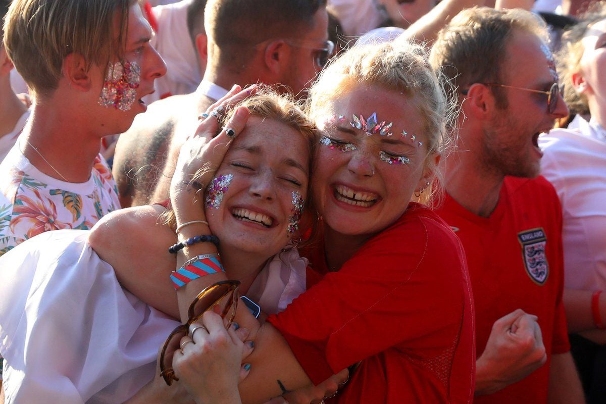 Two young women with glitter on their faces hug and smile joyfully in a crowd. One wears a red shirt with an England football crest; others around them also appear to be celebrating. Bright sunlight highlights the scene.