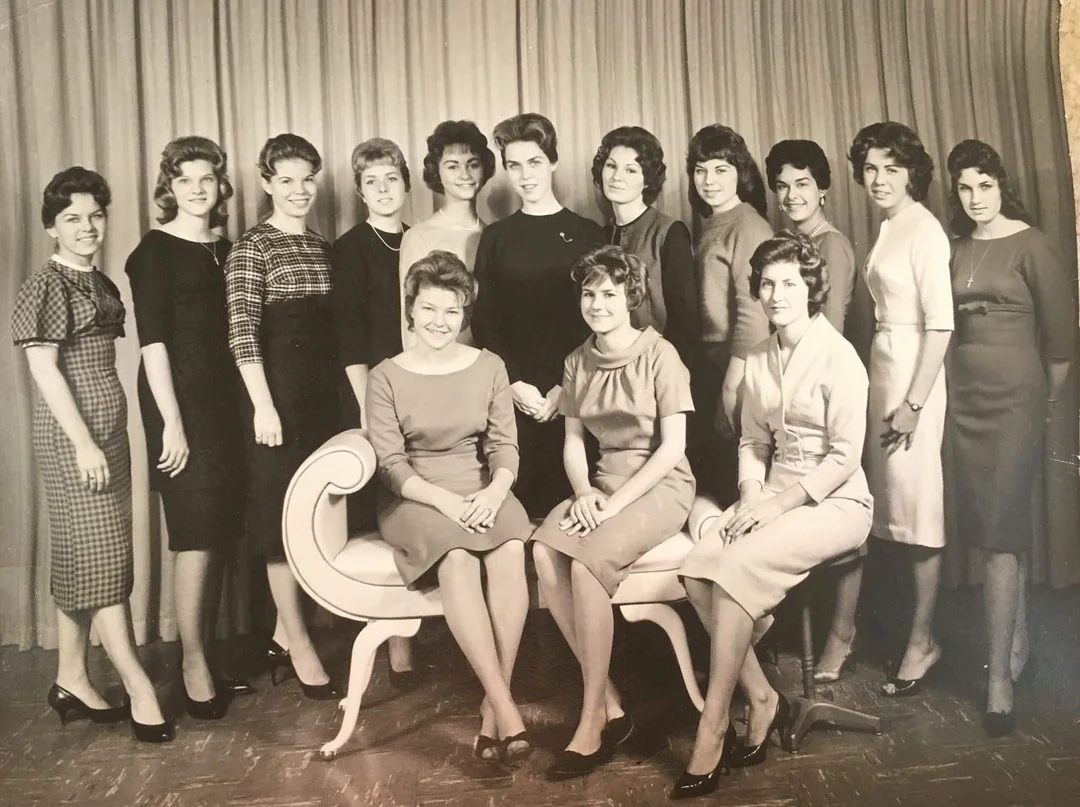 A black-and-white photo of fourteen women posing indoors, with two women seated on a cushioned bench and twelve women standing behind them. All are dressed in 1960s-style dresses and have neatly styled hair.