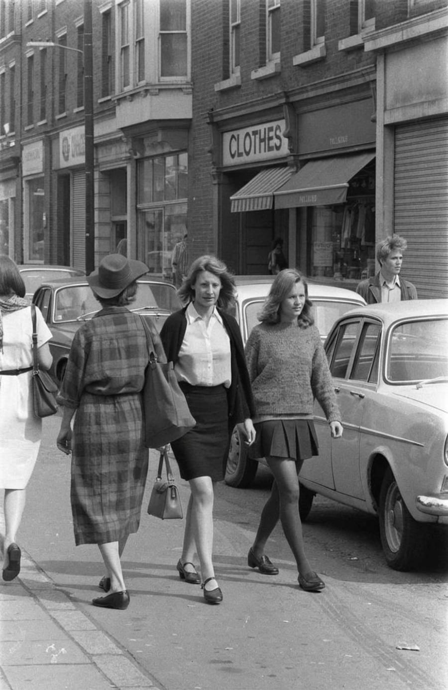 Black and white photo of people walking on a city sidewalk in front of parked cars and shops, including one with a striped awning labeled "CLOTHES." Two young women walk side by side in the foreground.