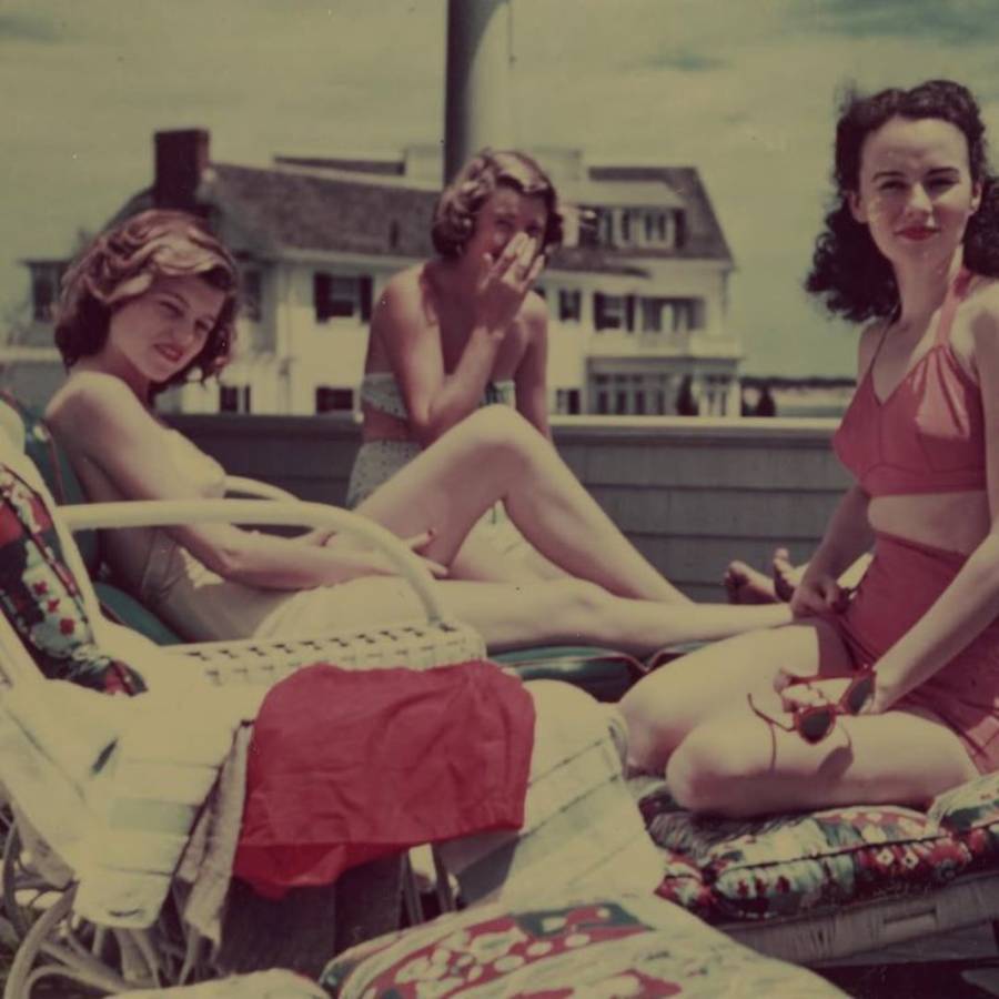 Three women in vintage swimsuits relax on lounge chairs outdoors, smiling and enjoying a sunny day. A red towel is draped over a chair, and houses are visible in the background under a blue sky.