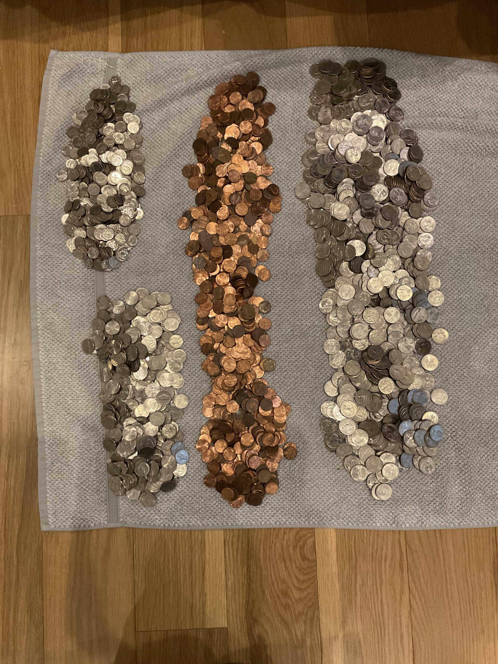 Four sorted piles of U.S. coins—nickels, dimes, pennies, and quarters—are arranged vertically on a towel placed on a wooden floor.