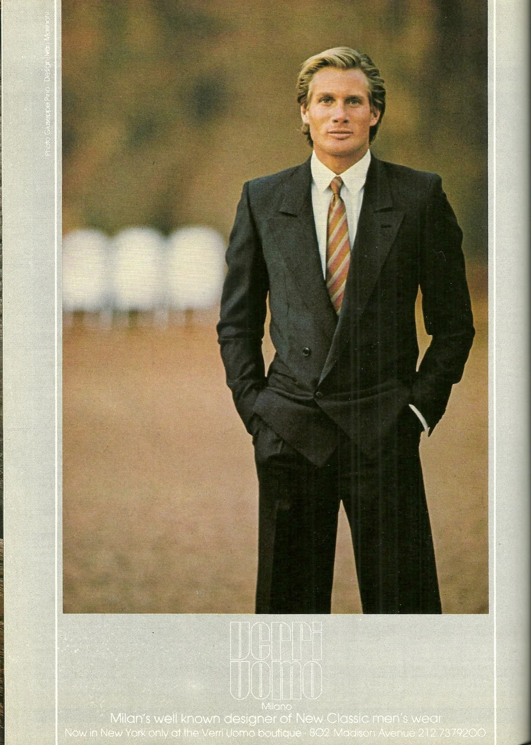A man in a black double-breasted suit with a striped tie stands outdoors with his hands in his pockets, looking at the camera. The background is blurred, showing a brown and green landscape.