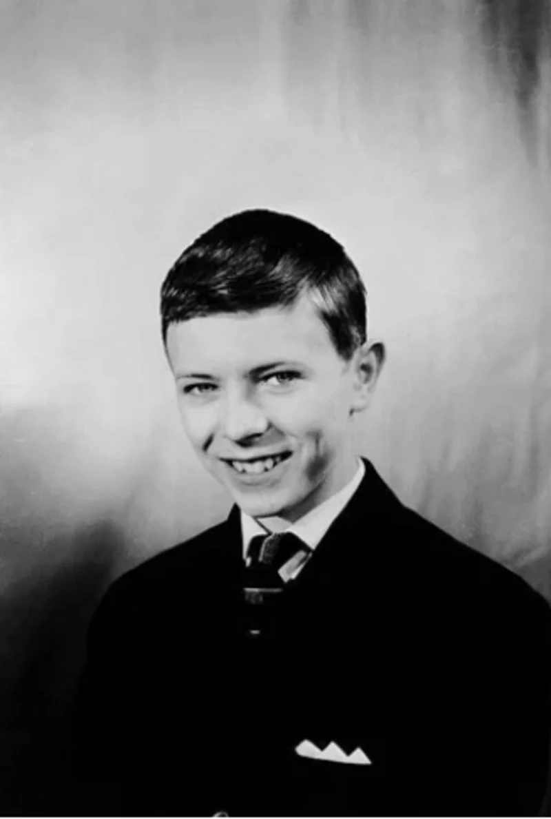 A young boy in a suit and tie smiles at the camera against a plain, light-colored background.