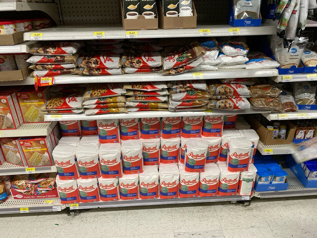 Shelves in a grocery store stocked with bags of Redpath sugar on the lower shelf and various types of cookies, crackers, and sugar products on the shelves above. The floor is white tile.