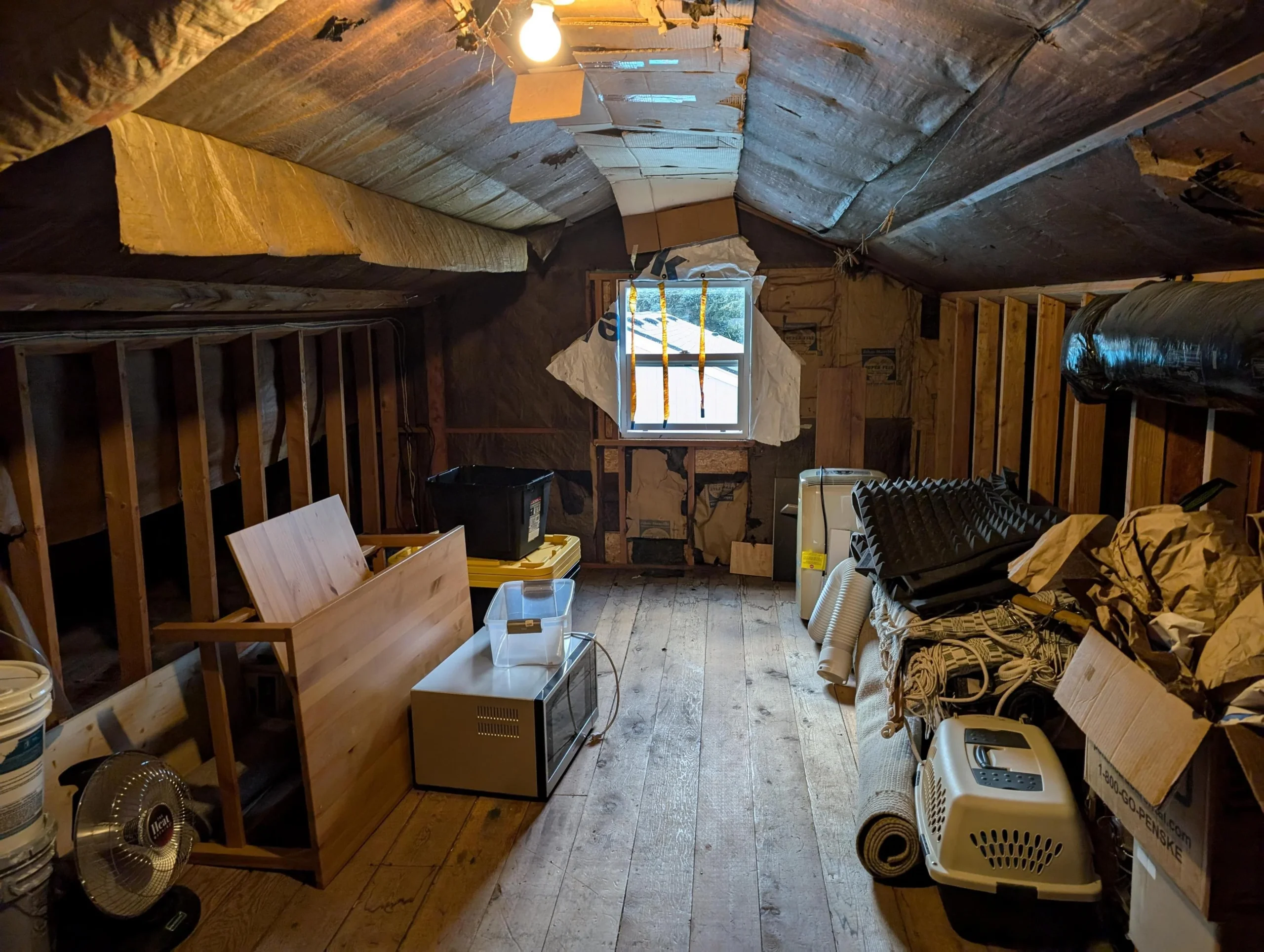 An unfinished attic with exposed insulation and wooden beams, cluttered with boxes, a table, appliances, and miscellaneous items, lit by a single bulb and natural light from a small window at the far end.