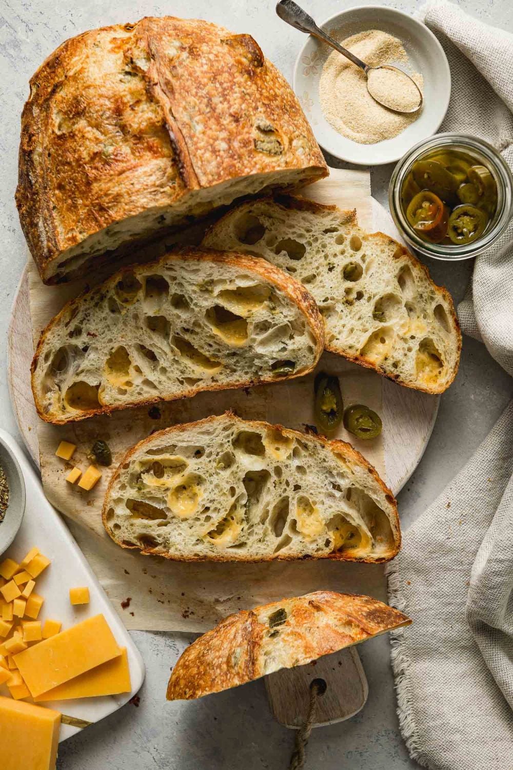 A rustic loaf of bread with cheddar cheese and jalapeño pieces is sliced on a round cutting board. Nearby are small bowls of jalapeños, shredded cheese, flour, and a linen napkin.