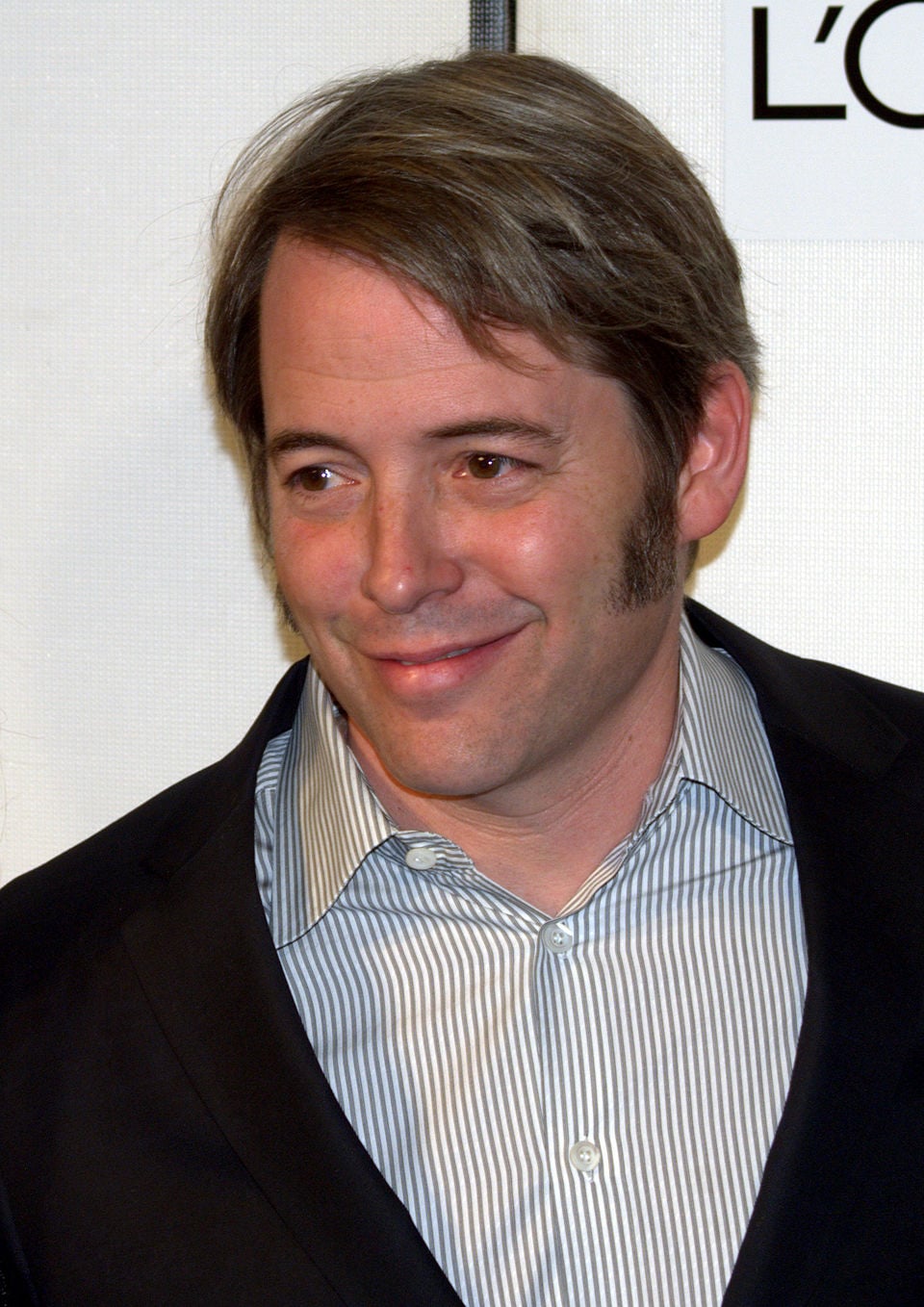 A man with short brown hair and sideburns is wearing a dark blazer over a light striped shirt. He is smiling and looking slightly to the side, standing in front of a white background with partial text visible.