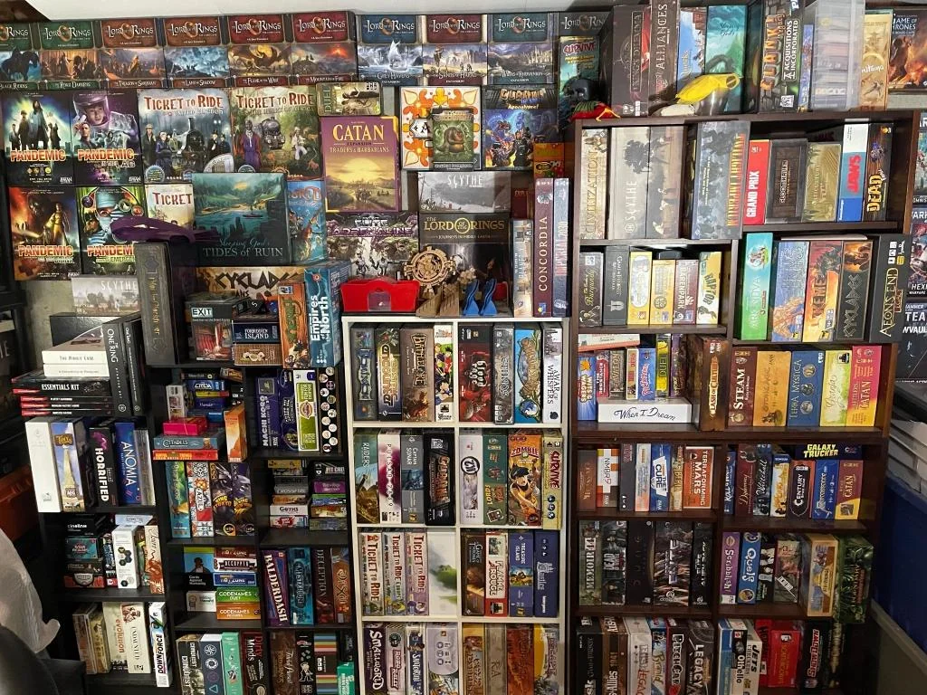 A large, crowded shelf filled with a wide variety of colorful board games, including titles like Ticket to Ride, Catan, and The Lord of the Rings, neatly organized and stacked closely together.