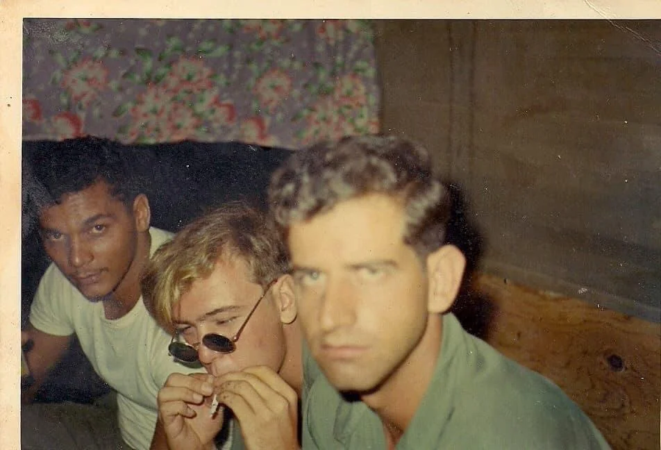 Three young men sit closely together indoors. One wears a white shirt and smiles slightly, the second adjusts his sunglasses and looks down, and the third, in focus at the front, gazes seriously at the camera. A floral curtain hangs behind them.