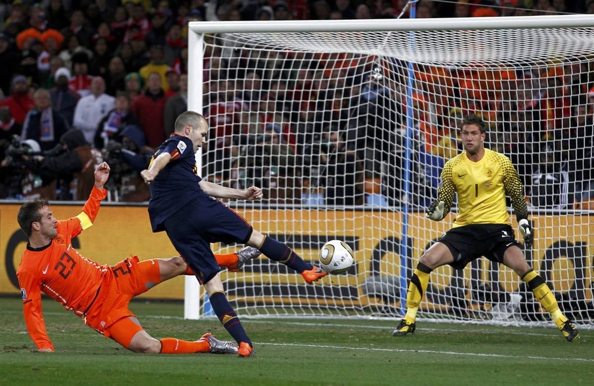 A soccer player in a dark blue uniform kicks a ball toward the goal as a player in an orange uniform slides to defend and the goalkeeper prepares to block during a crowded stadium match.