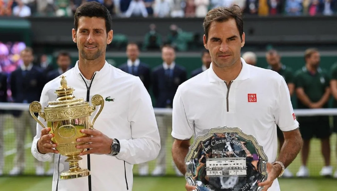 Two male tennis players stand side by side; one holds a gold trophy and the other holds a silver plate. They pose on a tennis court with people and officials in the background.