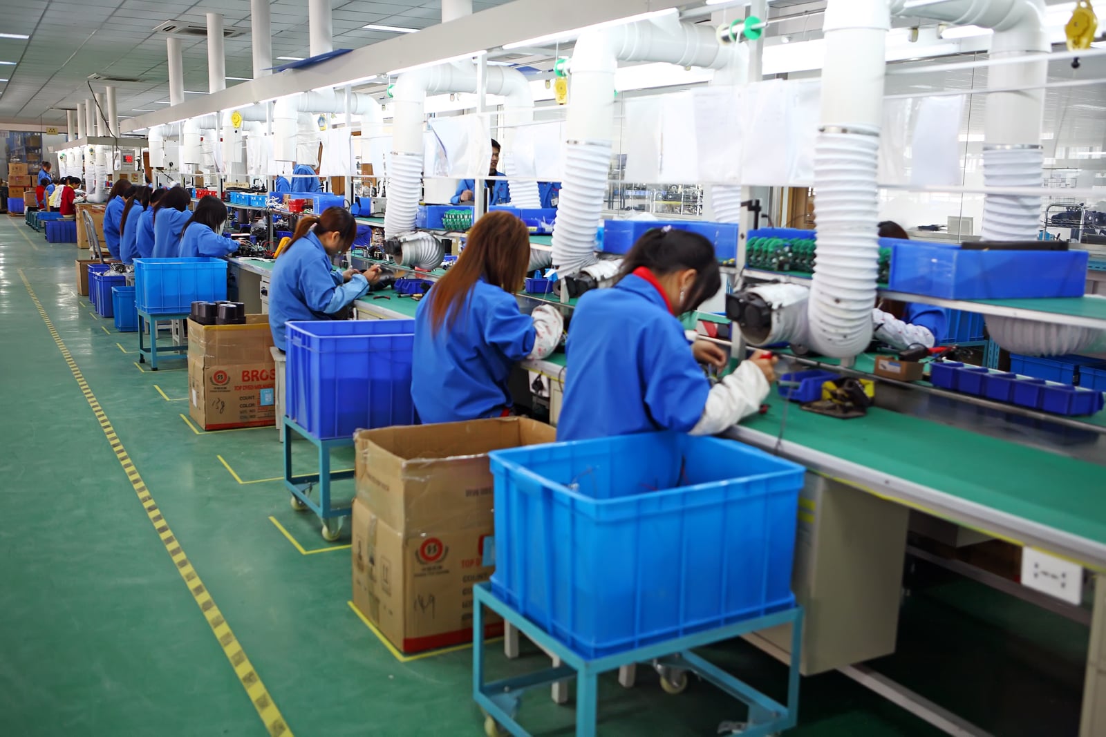 Workers in blue uniforms are seated in a row at a factory assembly line, handling electronic components with blue bins and cardboard boxes beside them in a brightly lit industrial setting.