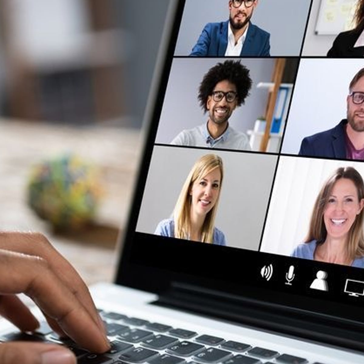 A person types on a laptop during a video conference call, with six people visible on the screen, each in their own separate window, smiling and engaging in the virtual meeting.