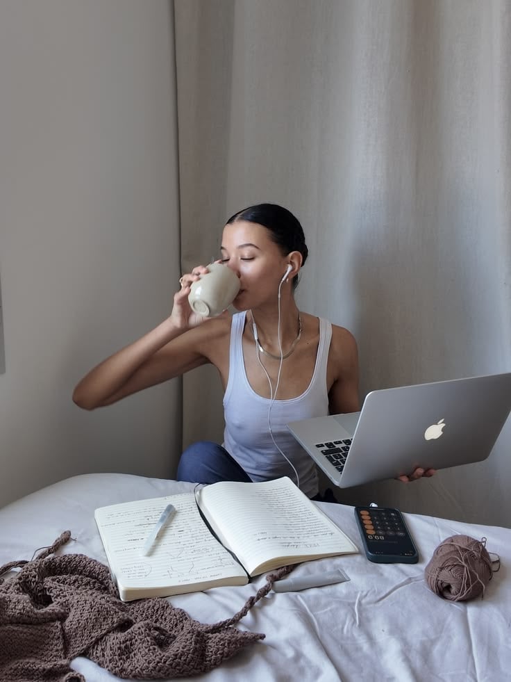A woman sits on a bed with a laptop on her lap, drinking from a mug. She wears earphones and a white tank top. Open notebooks, a pen, a calculator, and a ball of yarn are on the bed beside her.
