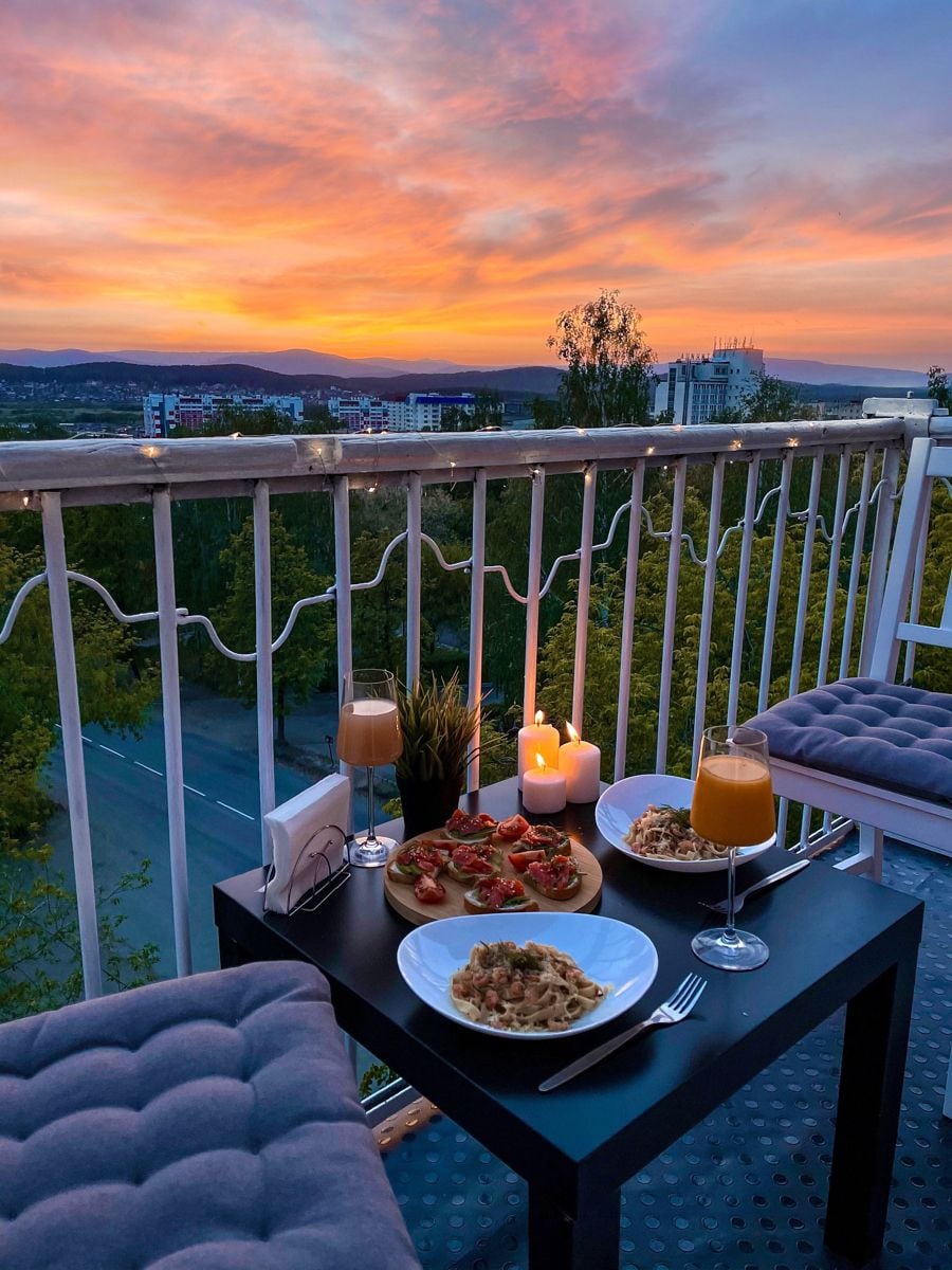 A cozy outdoor dinner setup on a balcony at sunset, featuring two plates of pasta, bruschetta, candles, two glasses of orange drink, and a scenic city and landscape view in the background.