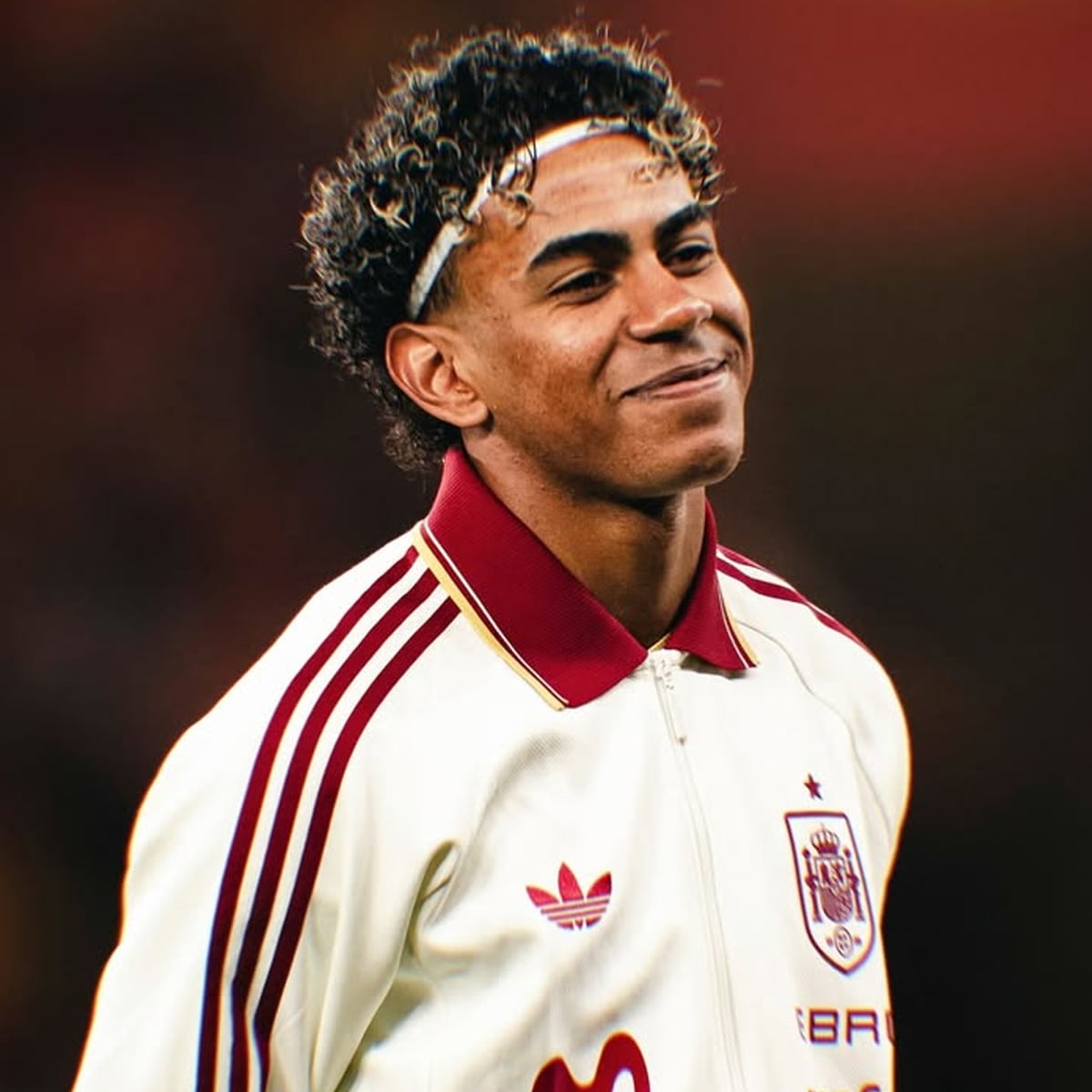 A young male soccer player with curly hair smiles while wearing a white Spain national team jacket with red stripes and logos, standing against a blurred background.