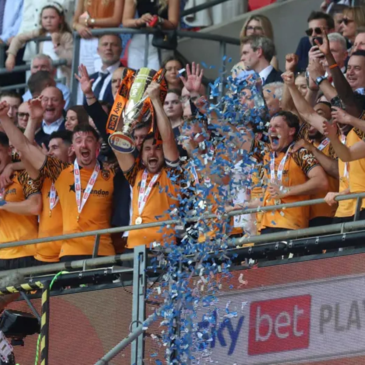 A jubilant soccer team in yellow and black jerseys celebrate with a trophy on a podium as blue confetti falls, surrounded by cheering teammates and spectators in formal attire.