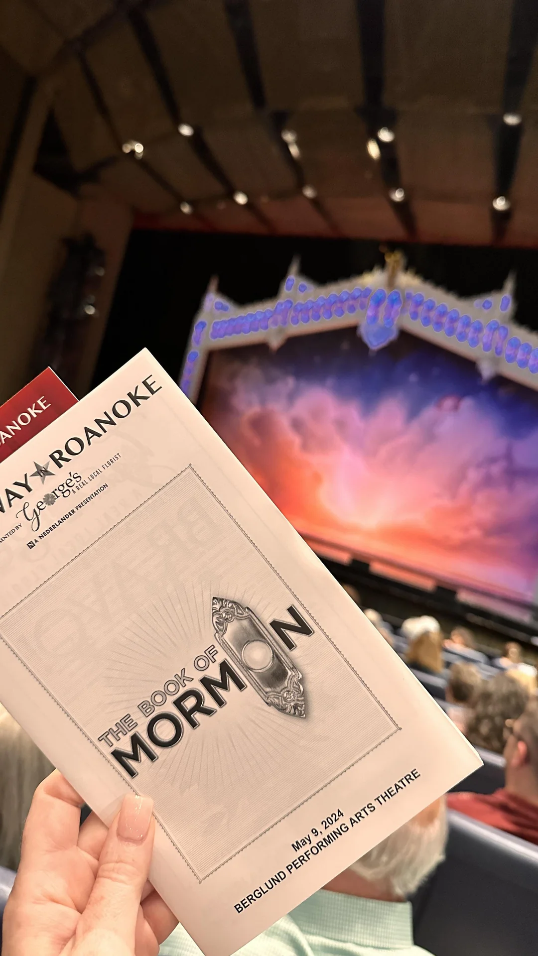 A hand holds a "The Book of Mormon" playbill in a theater, with an elaborate stage and audience seating visible in the background at the Berglund Performing Arts Theatre.