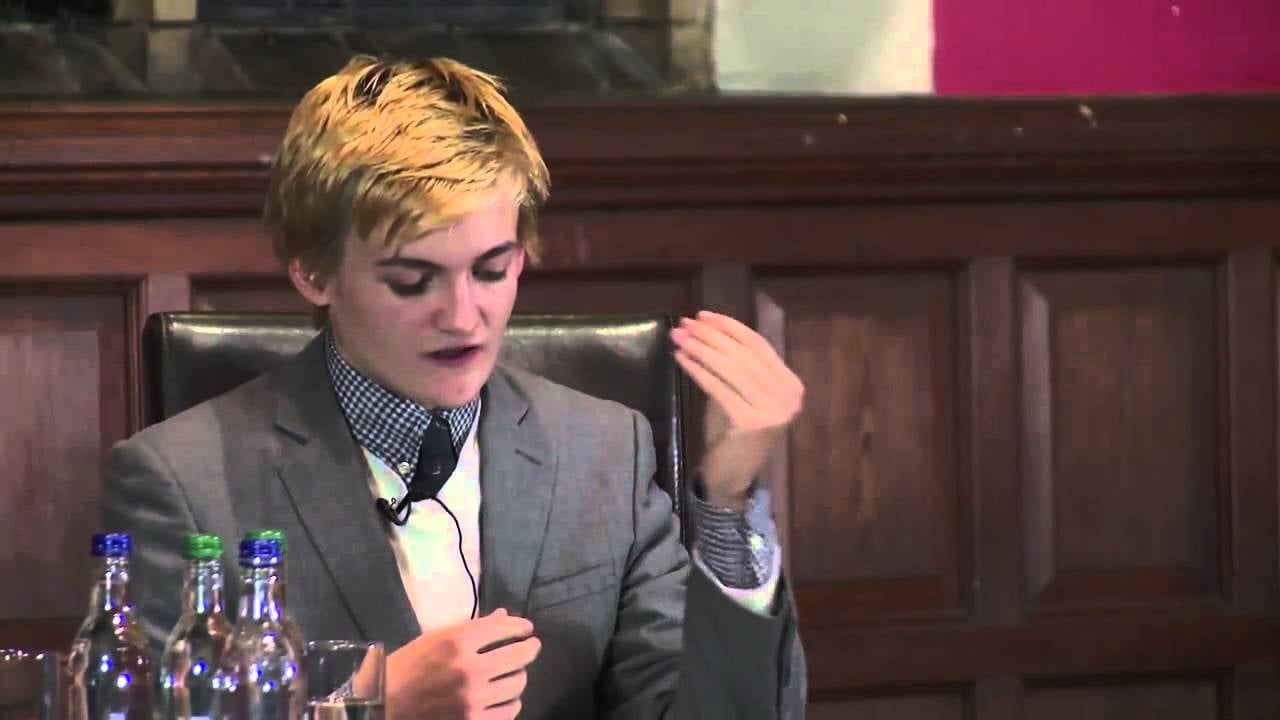 A person with short blonde hair, wearing a gray suit and patterned shirt, is seated at a table speaking and gesturing with their hand. Three water bottles are in front of them, and wood paneling is in the background.
