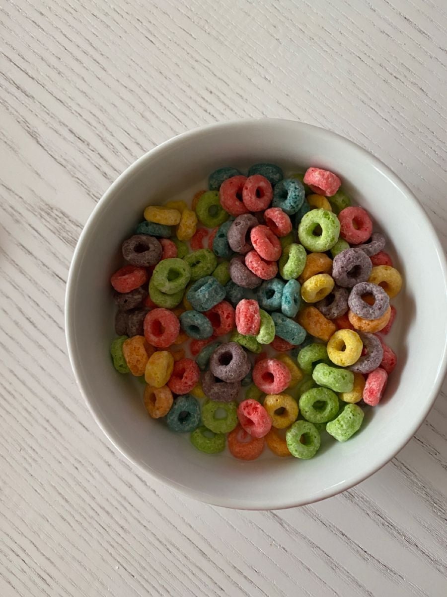 A white bowl filled with colorful, ring-shaped cereal sits on a light wood-grain table. The cereal pieces are yellow, green, red, orange, purple, and blue.