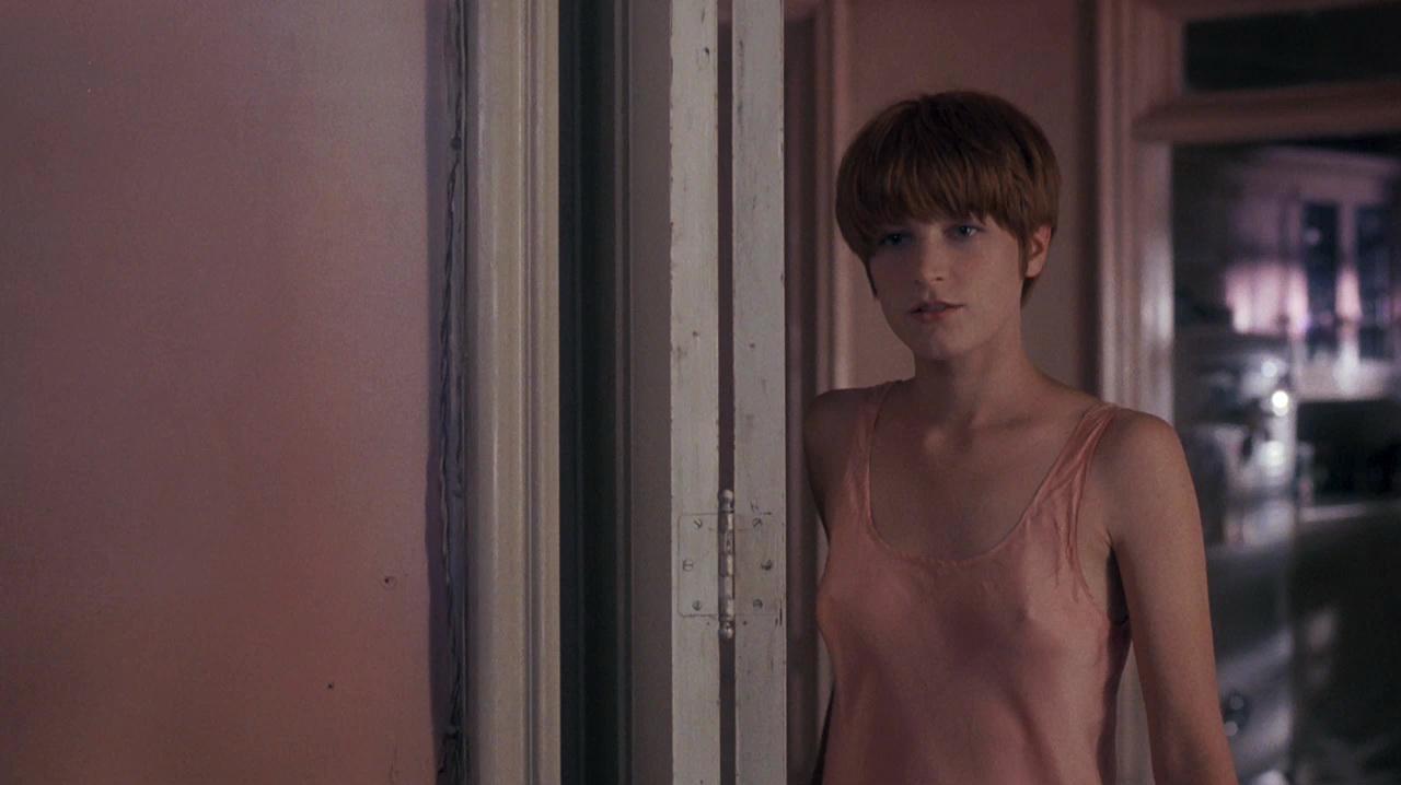 A young person with short, reddish hair stands in a doorway, wearing a light pink sleeveless top. The background is softly lit with pale pink and beige walls, creating a calm, introspective atmosphere.