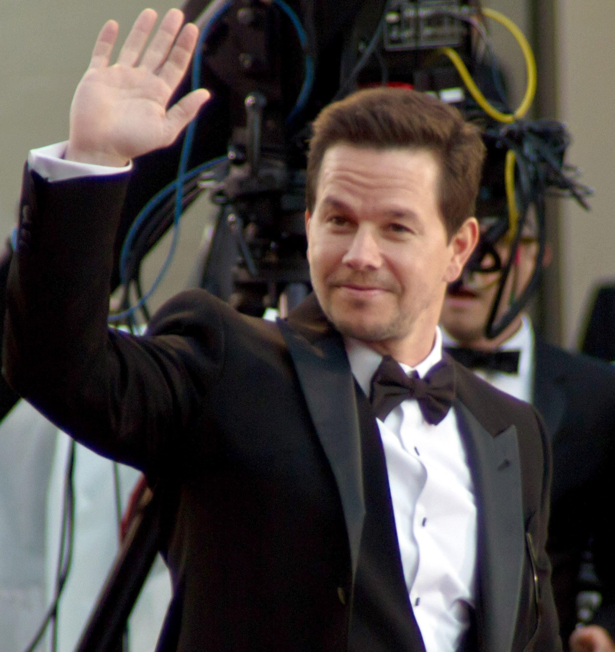 A man in a black tuxedo and bow tie smiles and waves with his right hand at an event, with camera equipment and cords visible in the background.