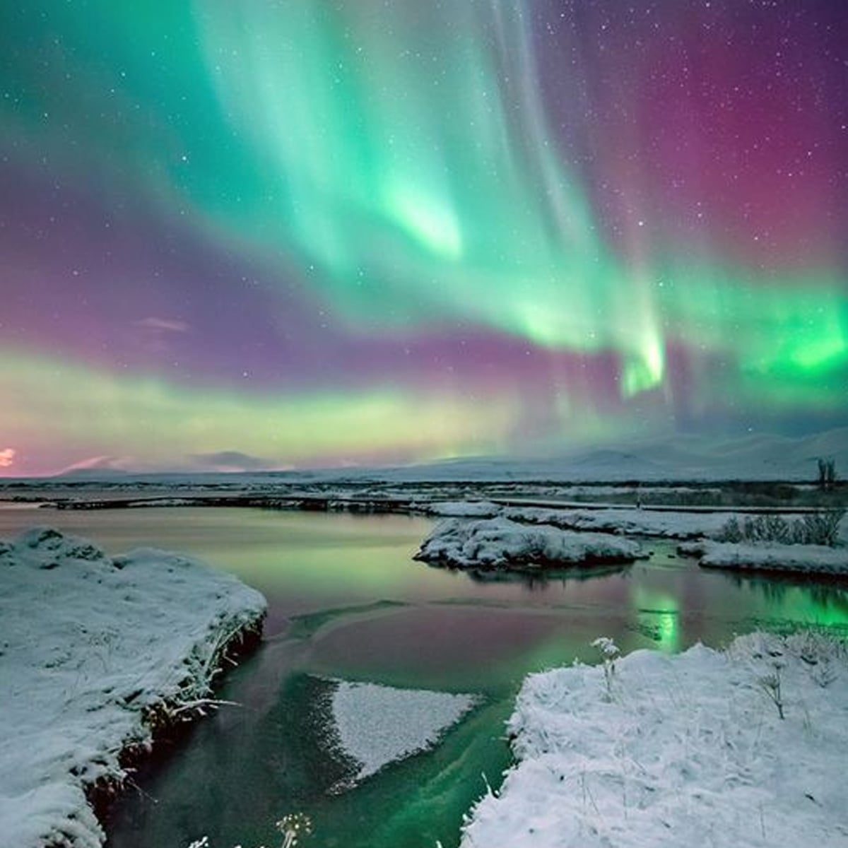 Colorful northern lights illuminate a snowy landscape with a river reflecting green and purple hues in the night sky; snow-covered banks and distant mountains complete the serene winter scene.