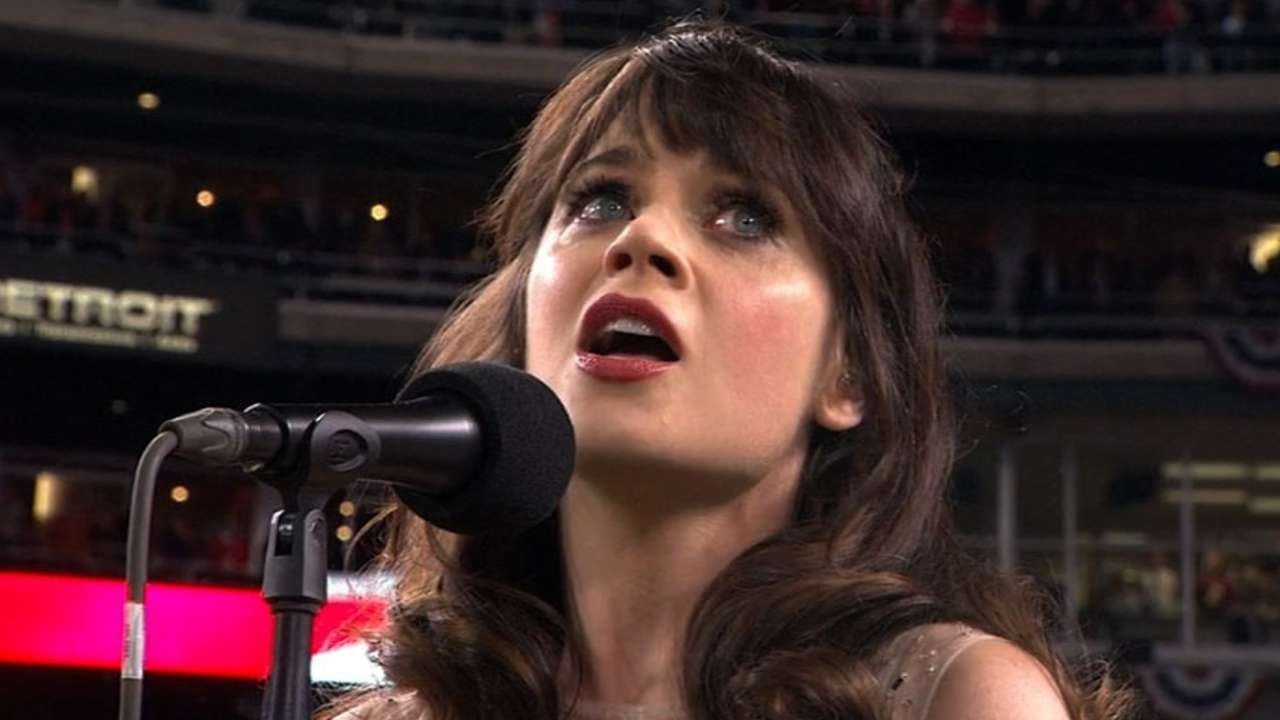 A woman with long brown hair stands in front of a microphone, looking upward with an expressive face. She appears to be singing at a stadium filled with an audience in the background.
