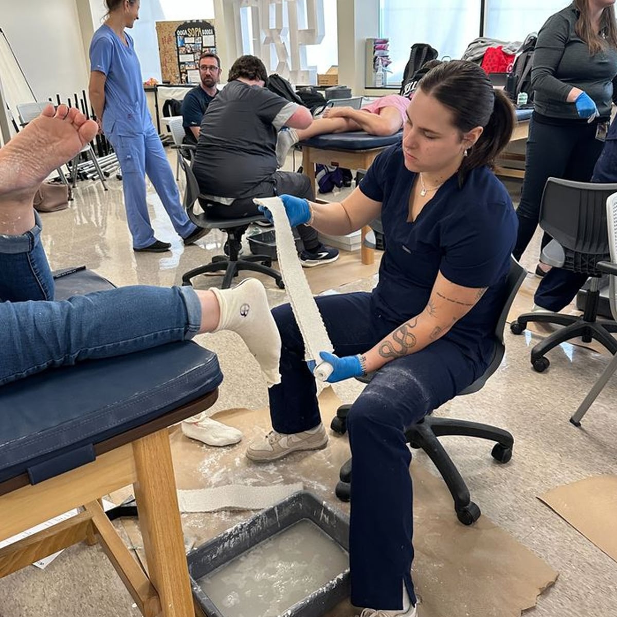 A woman in navy scrubs wraps a patient's foot in plaster bandages, possibly making a cast, in a clinical classroom setting with other people in the background practicing similar activities.