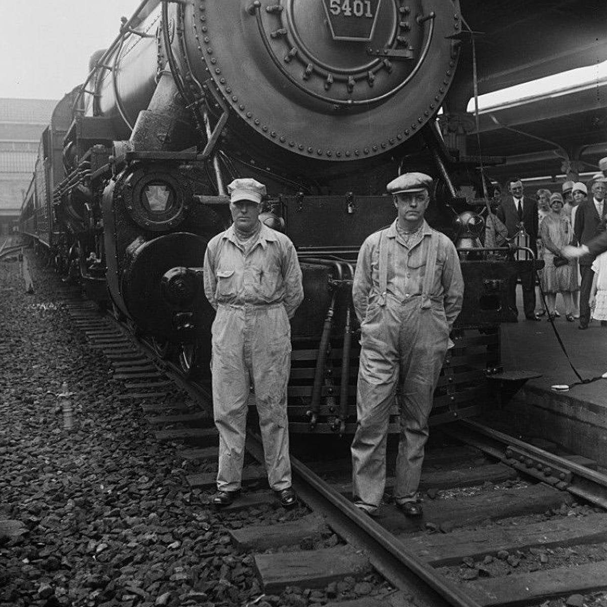 Two men in work overalls and caps stand on railroad tracks in front of a large steam locomotive at a train station, with people and a platform visible in the background.