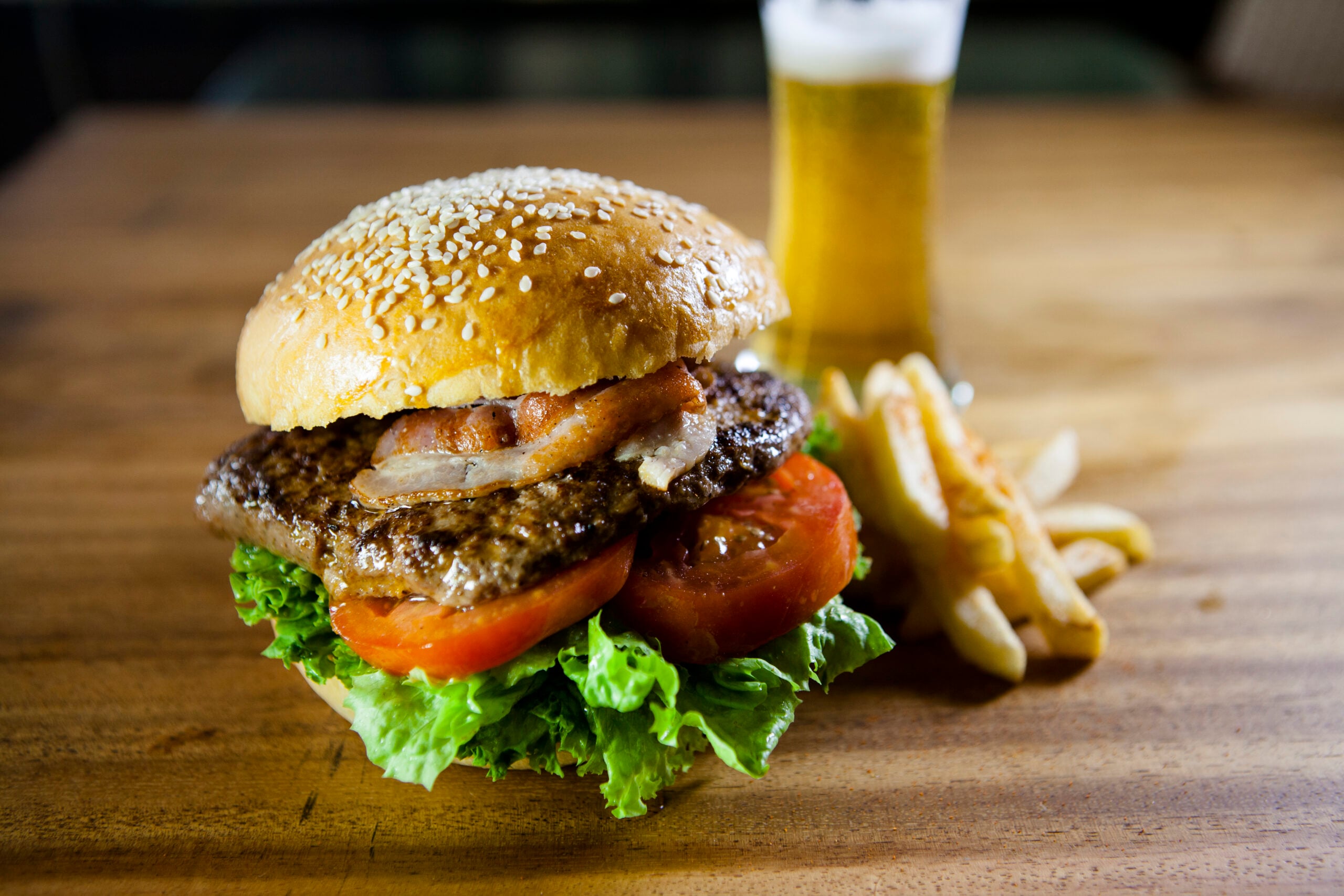 A cheeseburger with lettuce, tomato, bacon, and a sesame seed bun sits on a wooden table next to a small pile of French fries and a glass of beer in the background.