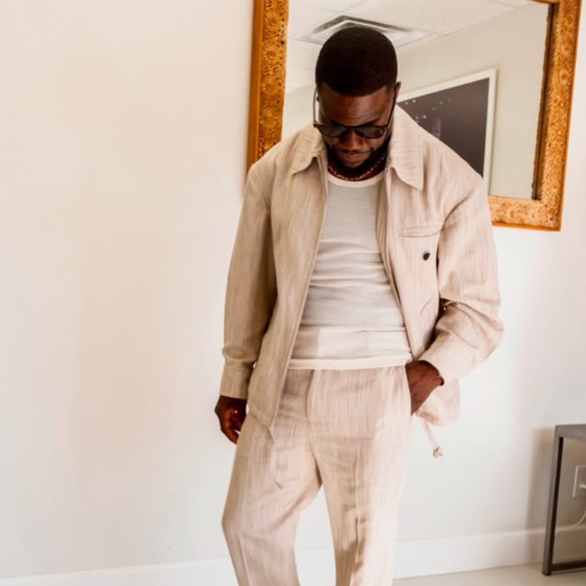 A man wearing sunglasses, a light beige suit, and a white shirt stands indoors, looking down with one hand in his pocket. A large ornate mirror hangs on the wall behind him.