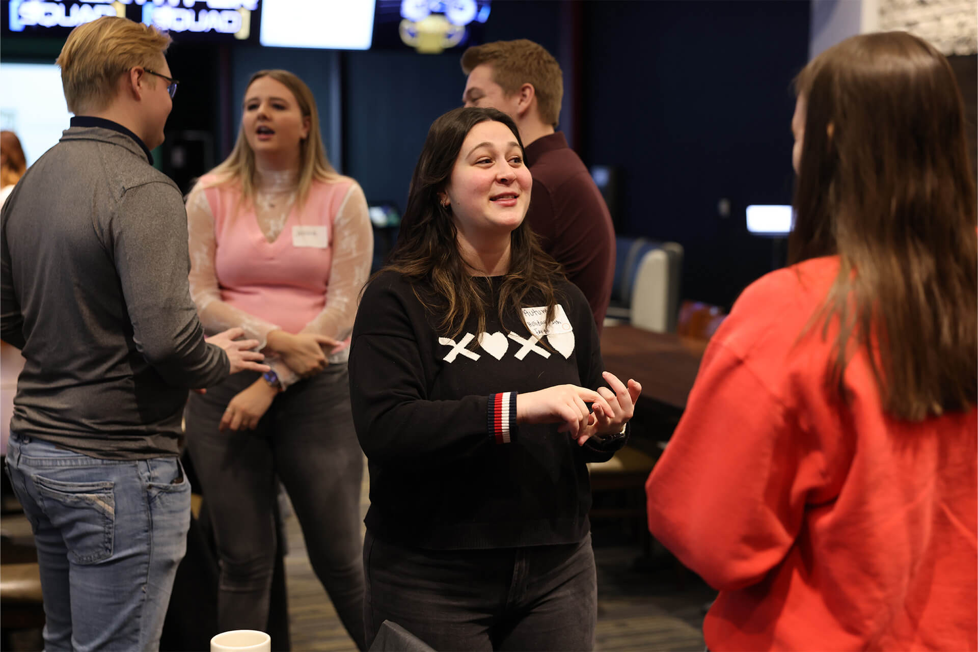 A group of young adults stand in a circle indoors, talking and laughing. One woman in a black sweater gestures while speaking to another woman in a red sweatshirt. Others in the background are also engaged in conversation.