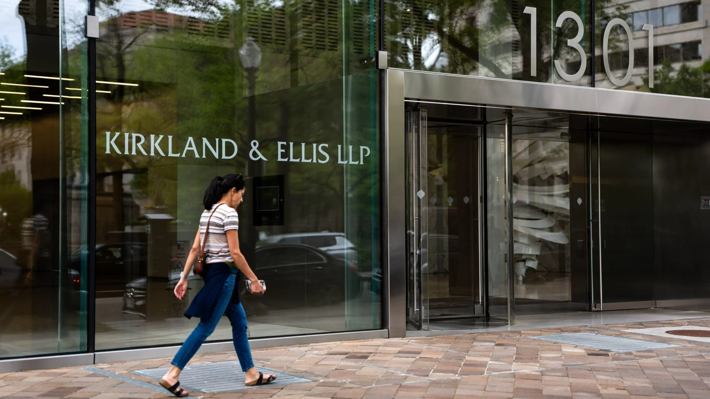 A woman walks past the glass entrance of the Kirkland & Ellis LLP office building at 1301, with reflections of trees and city life visible on the windows.