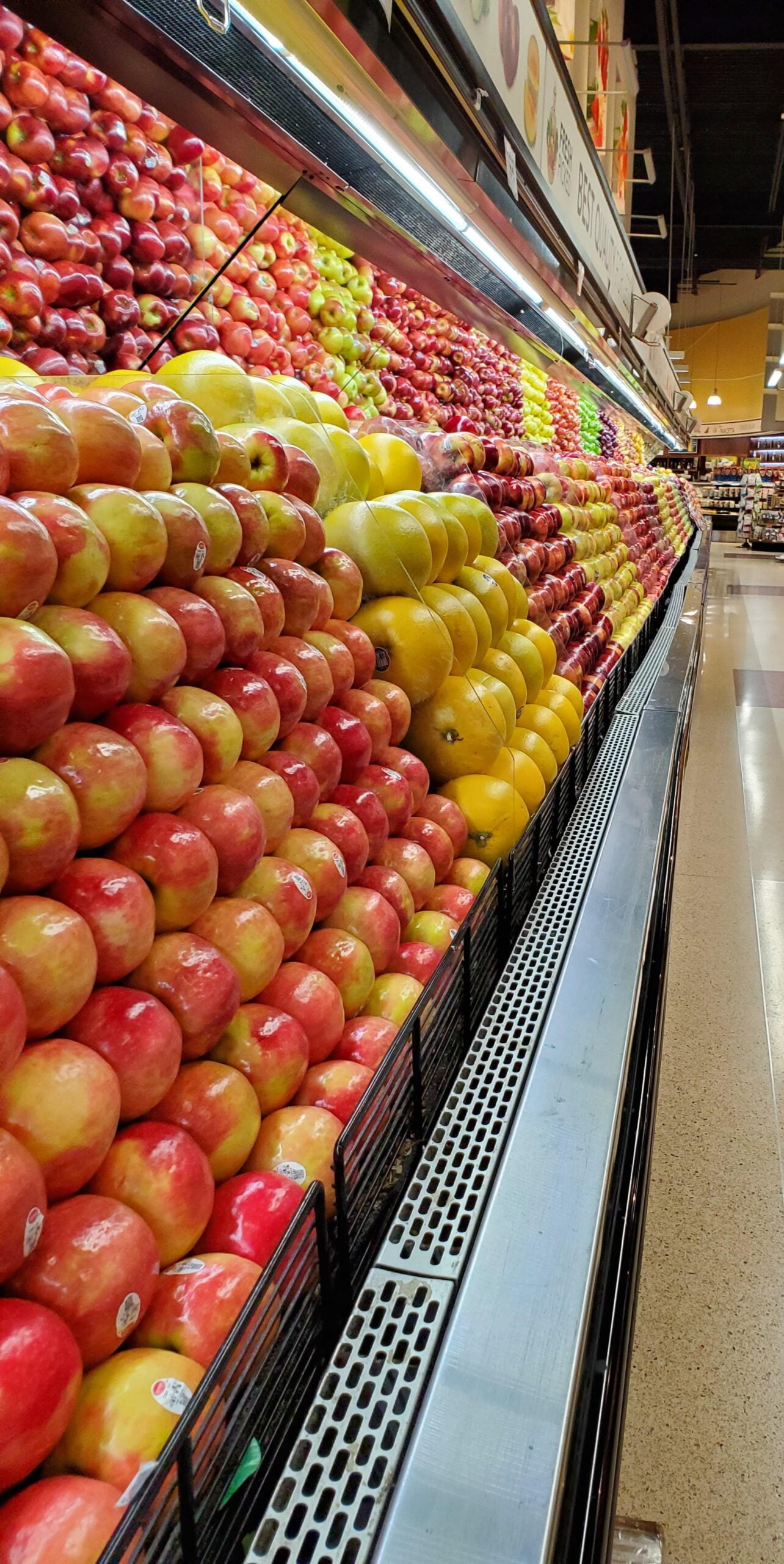 Rows of neatly stacked apples in various colors, including red, yellow, and green, fill a grocery store display. The apples are organized in diagonal lines along a long refrigerated shelf.