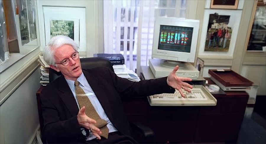 An older man in a suit and tie sits in an office gesturing with his hands. Behind him is a vintage computer with a colorful screen and a keyboard, surrounded by framed photos and documents on the wall.