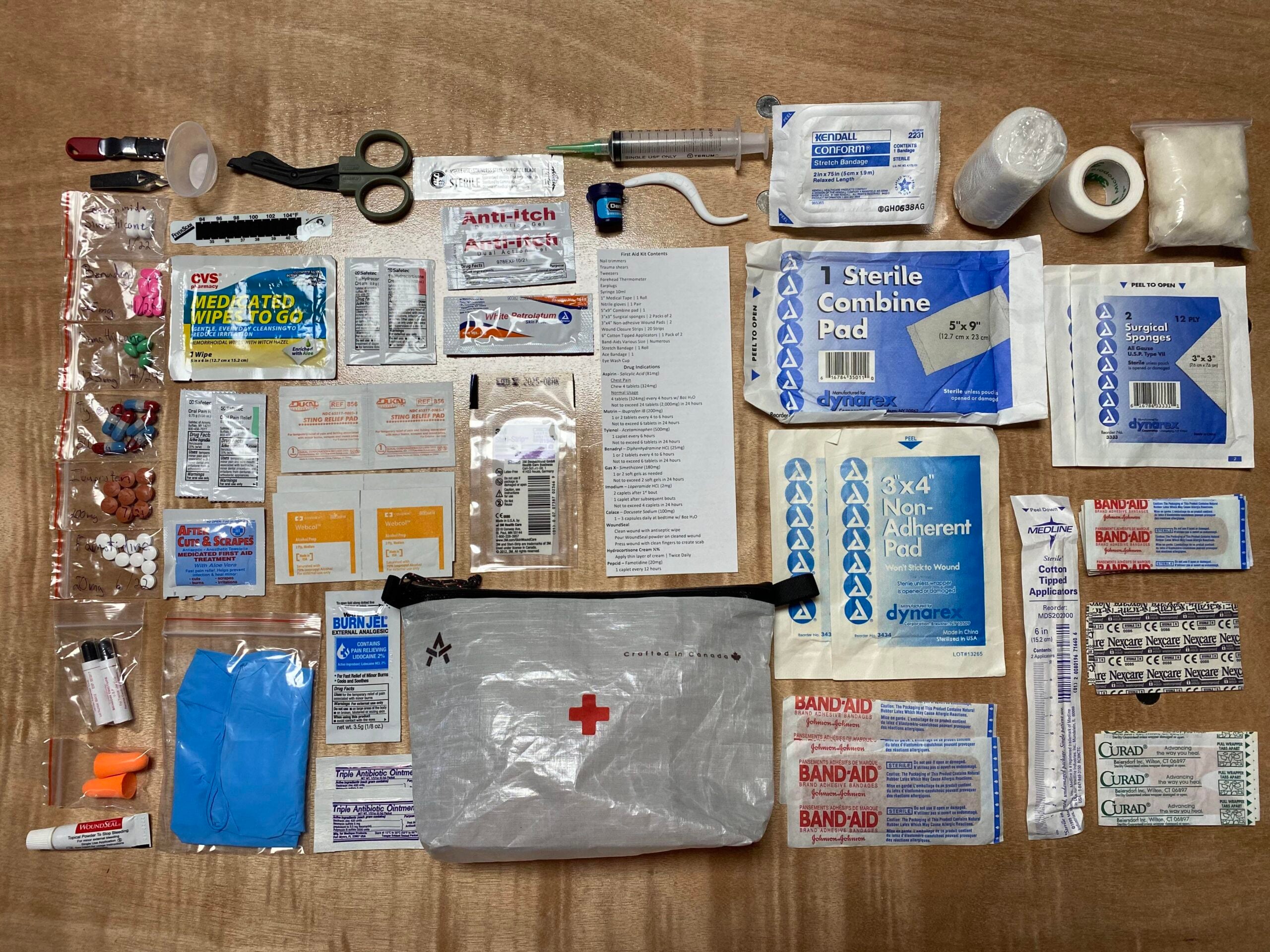 A neatly arranged first aid kit on a wooden surface, showing bandages, gauze pads, medications, ointments, medical tape, scissors, gloves, syringes, tweezers, and a clear pouch with a red cross symbol.