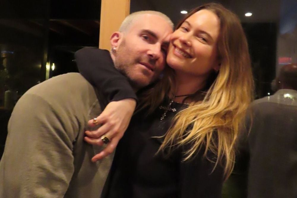 A woman with long blonde hair smiles and hugs a man with short light hair. The man wears a beige jacket and looks at the camera while the woman wraps her arm around his neck. They are indoors at night.