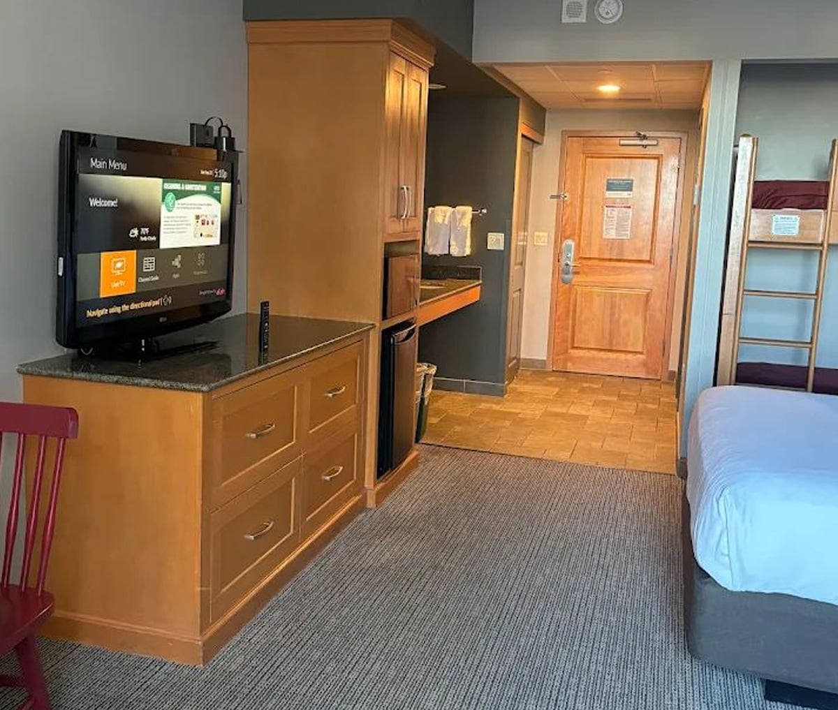 Hotel room interior with a wall-mounted TV on a dresser, wooden cabinets, mini fridge, microwave, and desk area. A bed is partially visible; bunk beds are near the door. Door and tiled entryway are in the background.