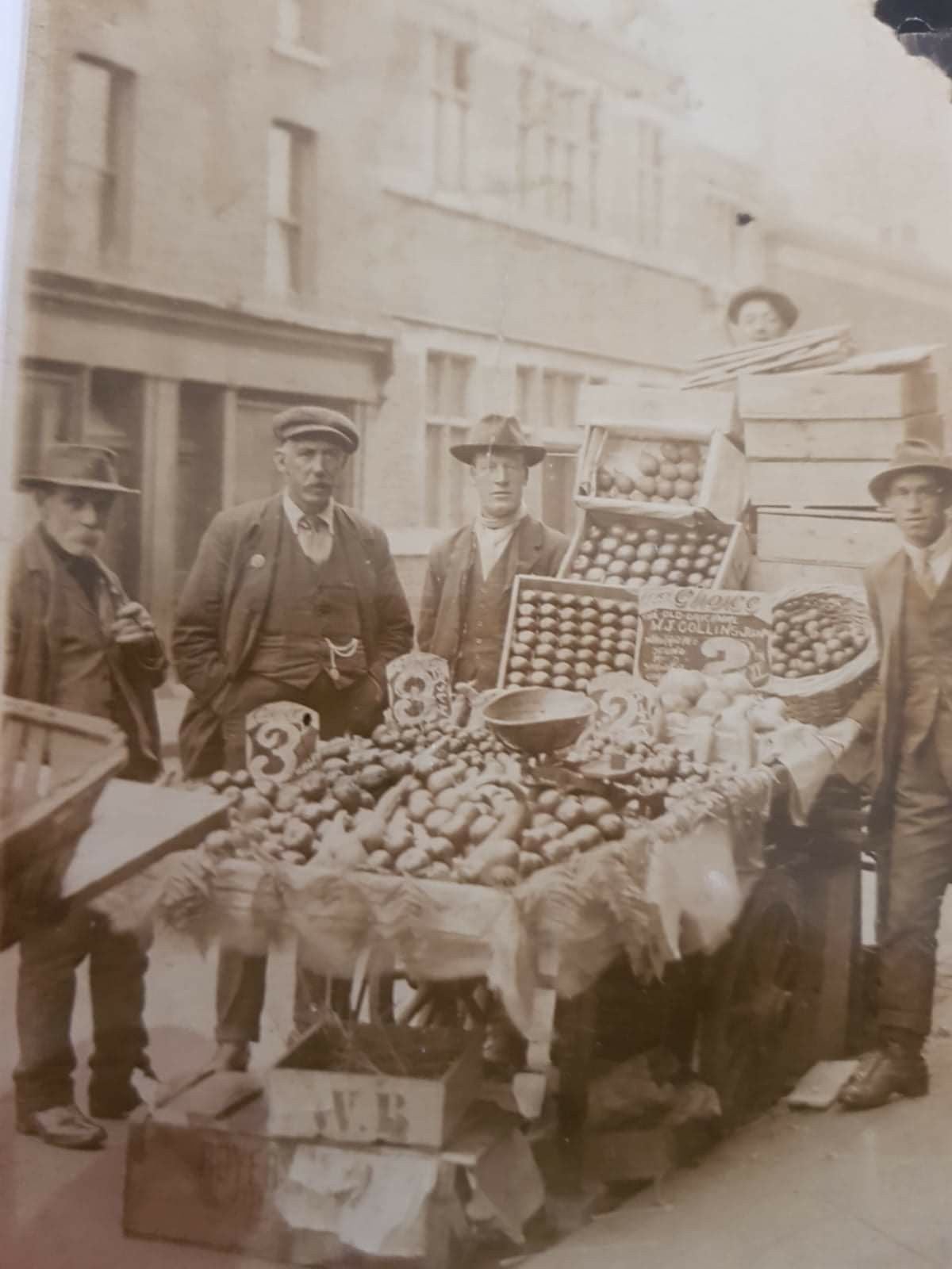A sepia-toned photo of five men in suits and hats standing beside a street fruit and vegetable stall, with produce like apples and eggs displayed in boxes and signs showing prices. Brick buildings are visible in the background.