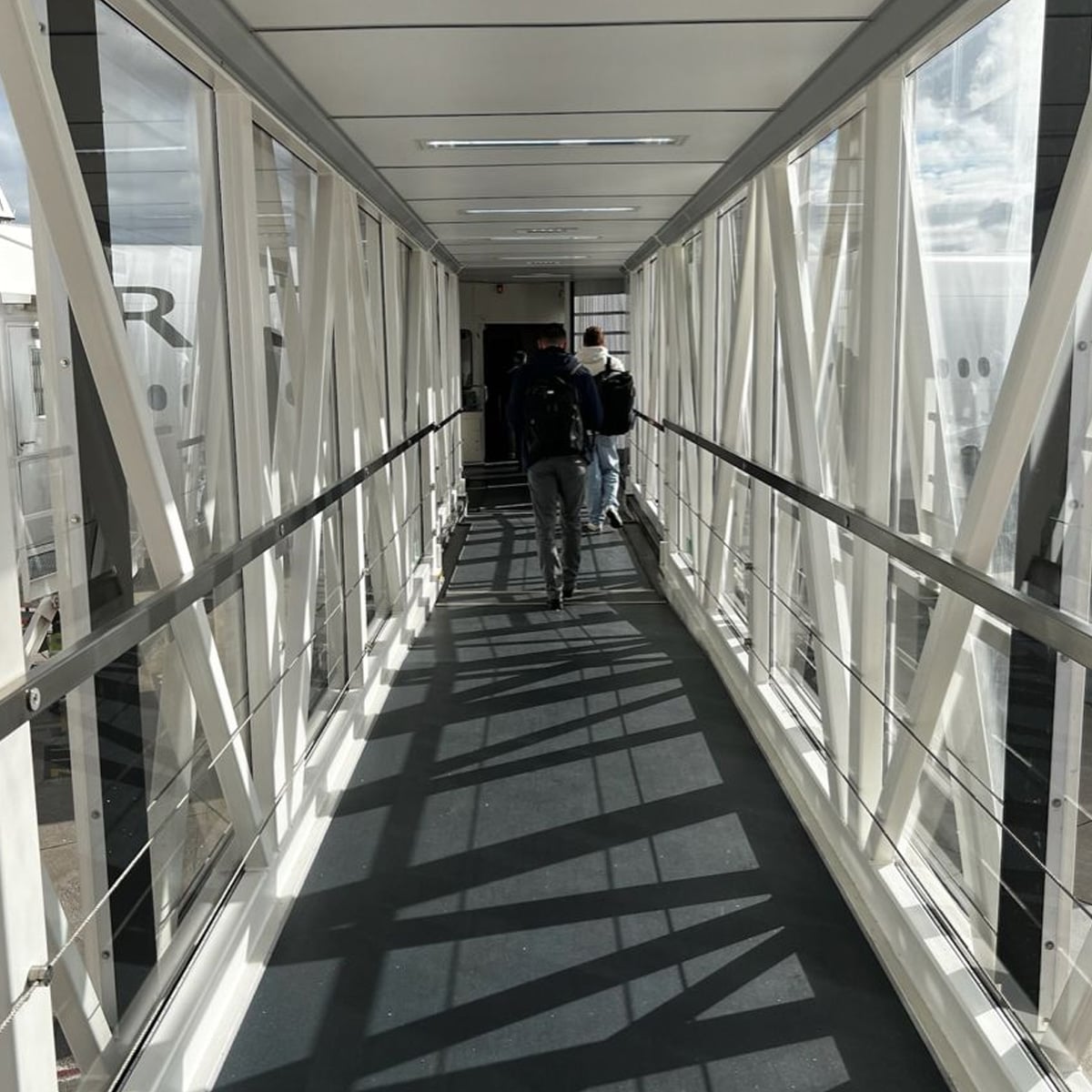 Two people walk down a glass-walled airport jet bridge toward an airplane, with sunlight casting shadows on the floor.