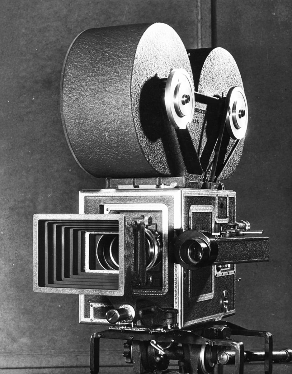 Black and white photo of a vintage film camera with two large reels on top, a square lens hood, and various mechanical controls, set against a plain background.