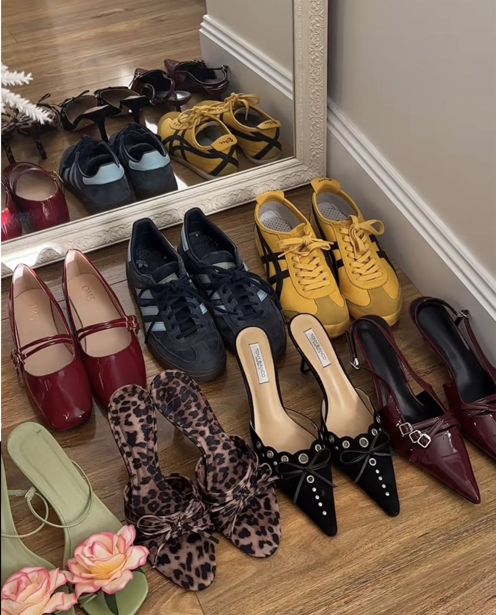 A variety of women's shoes, including red flats, black sneakers, yellow sneakers, green sandals with pink flowers, leopard print flats, and black pointed heels, are arranged neatly on a wooden floor in front of a mirror.