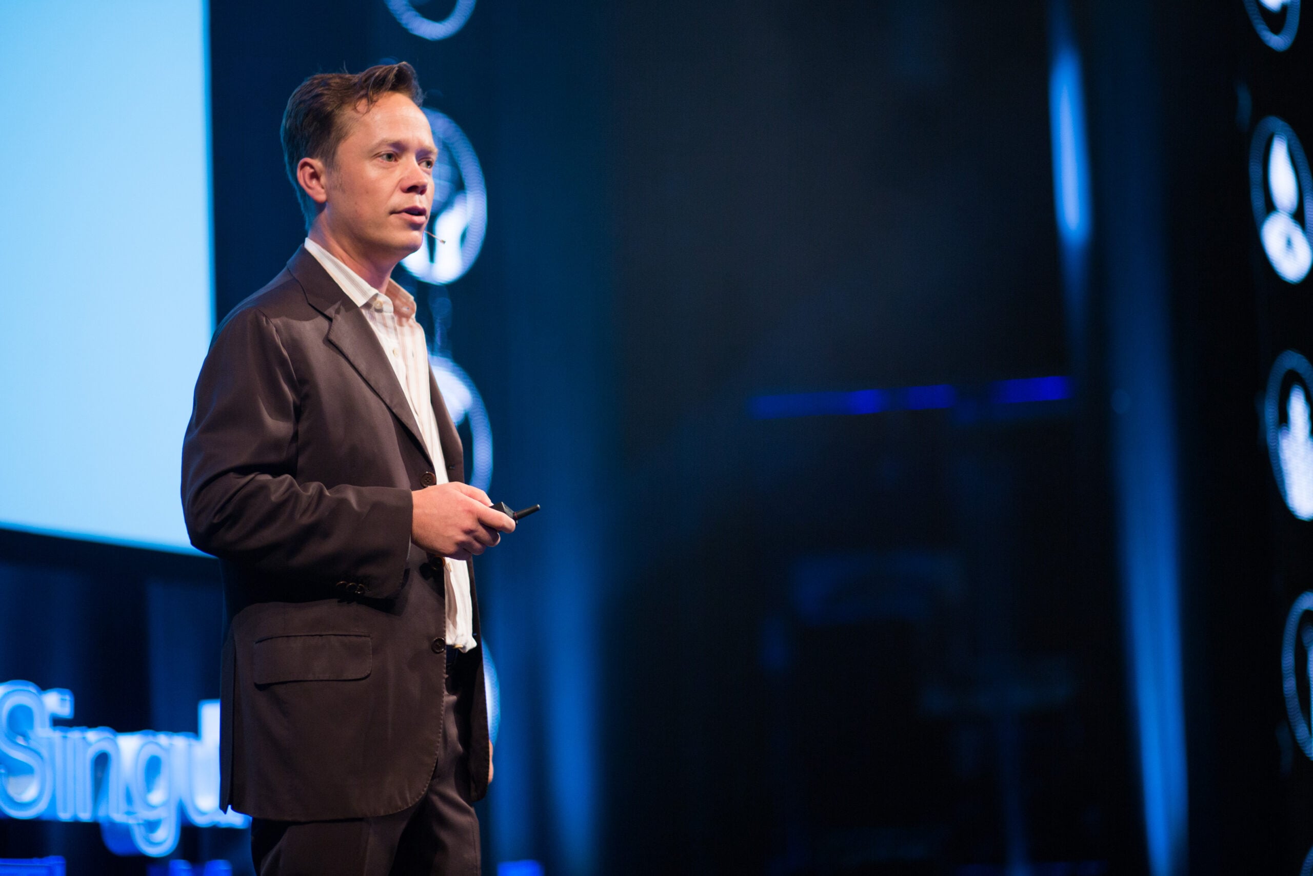 A man in a suit stands on stage holding a small device, speaking in front of a blue-lit background with a partial word "Singul" visible on a screen.