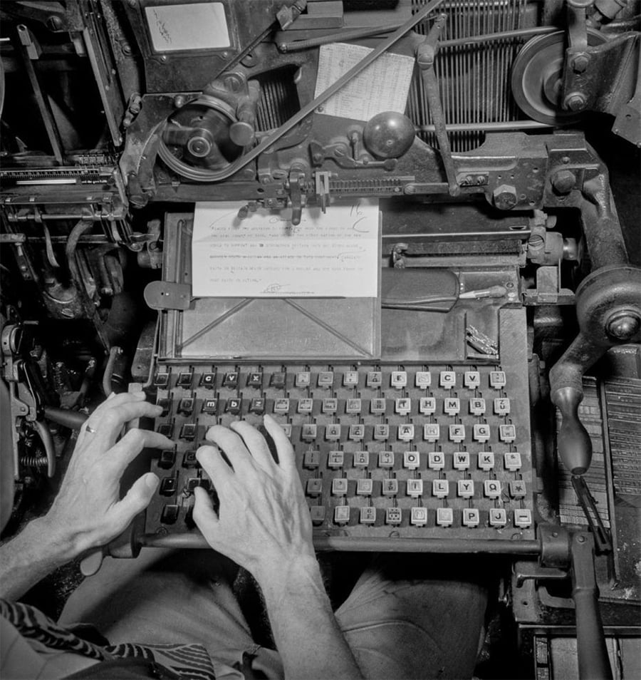 A person’s hands typing on a large, vintage Linotype machine keyboard, with mechanical parts and a sheet of paper visible above the keys. The image is black and white, showing detailed machinery and old printing technology.