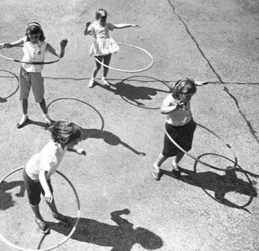 Five children play outside with hula hoops on a paved surface, casting long shadows in the sunlight. The image is black and white, giving it a nostalgic, vintage feel.